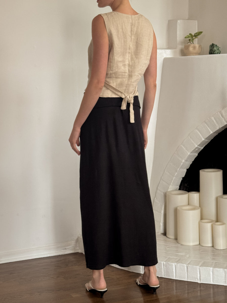 A woman shown from the back wearing a sleeveless beige top with a tie detail at the waist and a long black skirt, standing in a room with a white fireplace and candles.