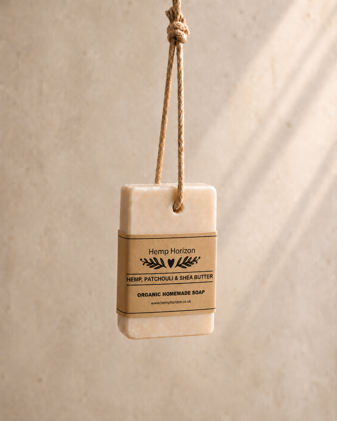 A rectangular bar of Hemp Horizon Hemp, Patchouli & Shea Butter organic homemade soap hangs from a natural jute rope tag against a textured beige wall with soft diagonal shadows. Hemp Horizon