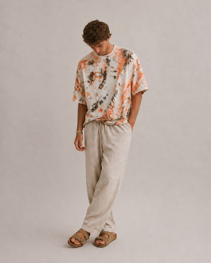 A man wearing a short-sleeve tie-dye t-shirt in orange, brown,  paired with light beige Hemp drawstring pants and brown sandals, standing against a neutral light gray studio background. Hemp Horizon