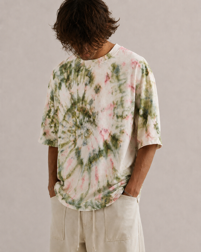 A person wearing an oversized short-sleeve tie-dye T-shirt with a spiral pattern in olive, pink, and beige tones, paired with light hemp beige pants, photographed against a plain light gray studio background. Hemp Horizon