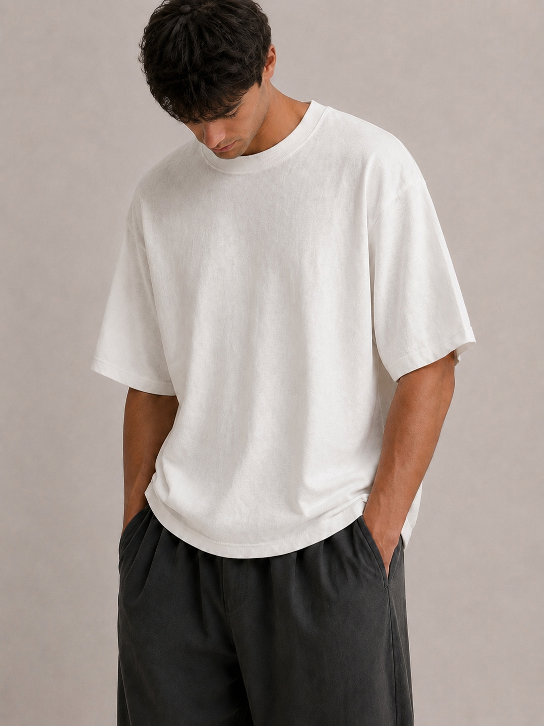 A man wearing an oversized white short-sleeve crewneck t-shirt and dark charcoal gray pants, standing against a neutral beige background. Hemp Horizon