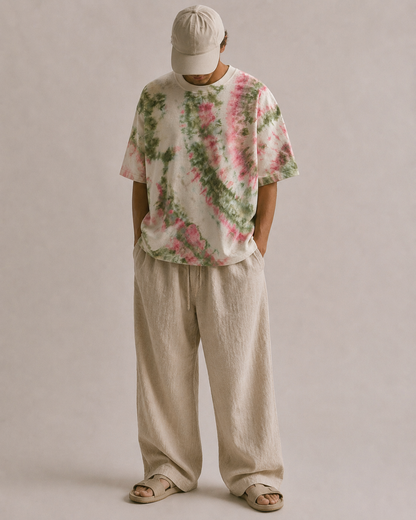 A person wearing a loose-fit short-sleeve T-shirt with a pink and green tie-dye pattern on an off-white base, paired with matching beige hemp pants, a beige baseball cap, and beige slide sandals, standing against a plain beige studio background. Hemp Horizon