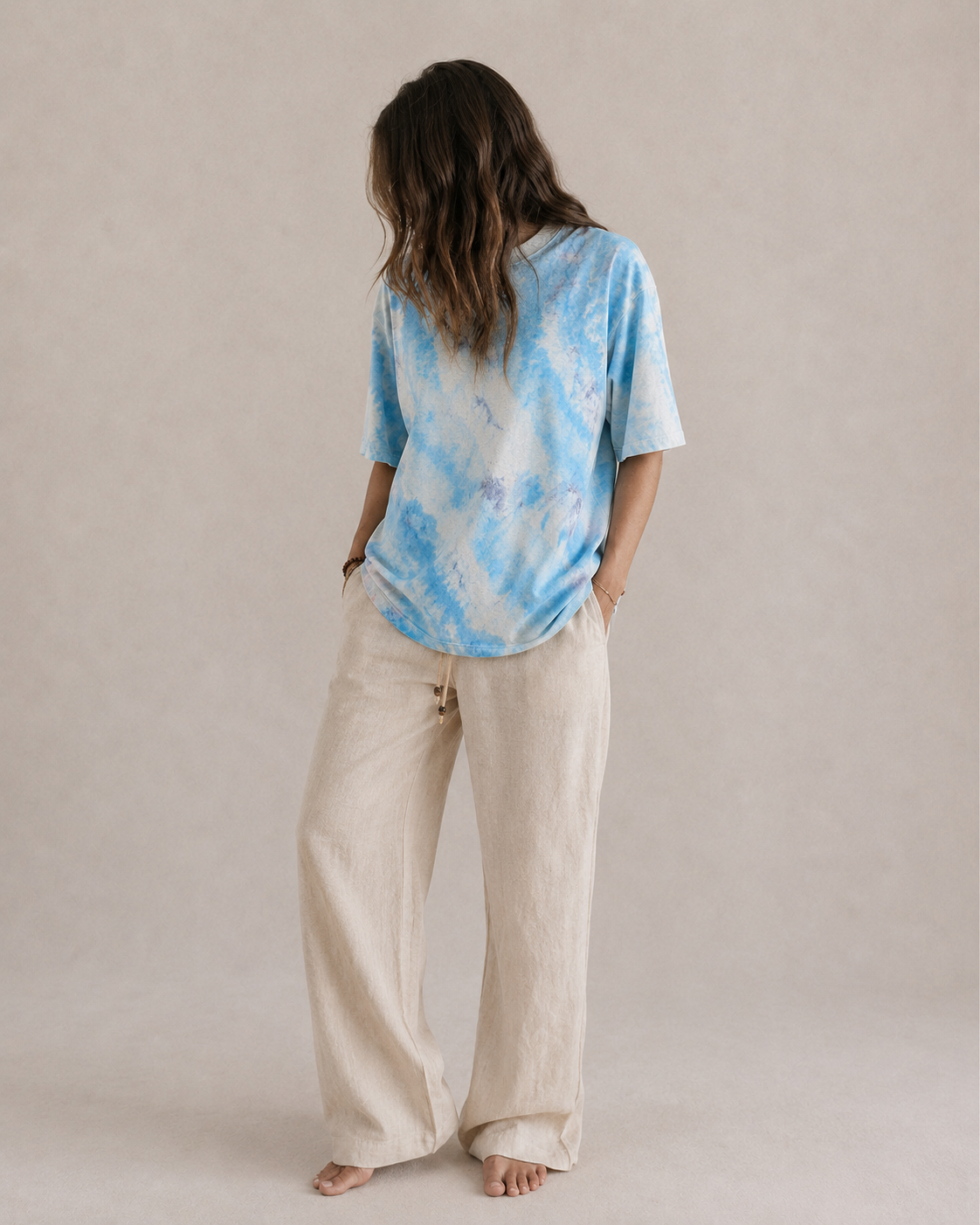 A woman wearing a light blue and navy and natural tie-dye t-shirt and beige wide-leg linen pants, standing against a neutral beige studio backdrop. Hemp Horizon